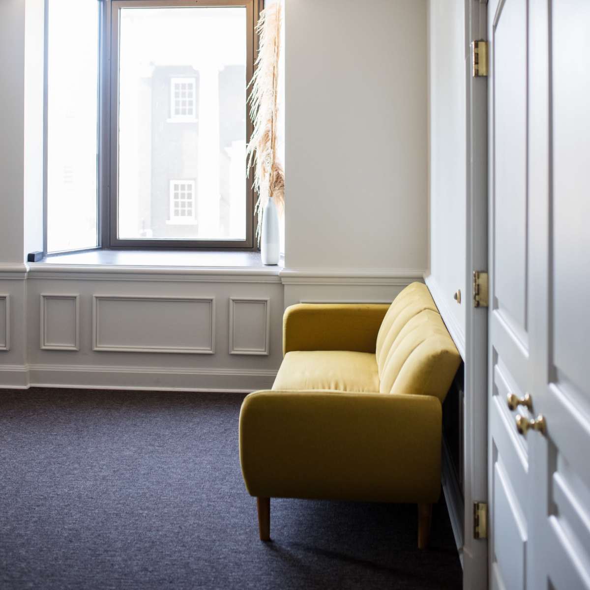 A yellow couch is positioned near a large window in a modern office space with light gray walls and a dark carpet.