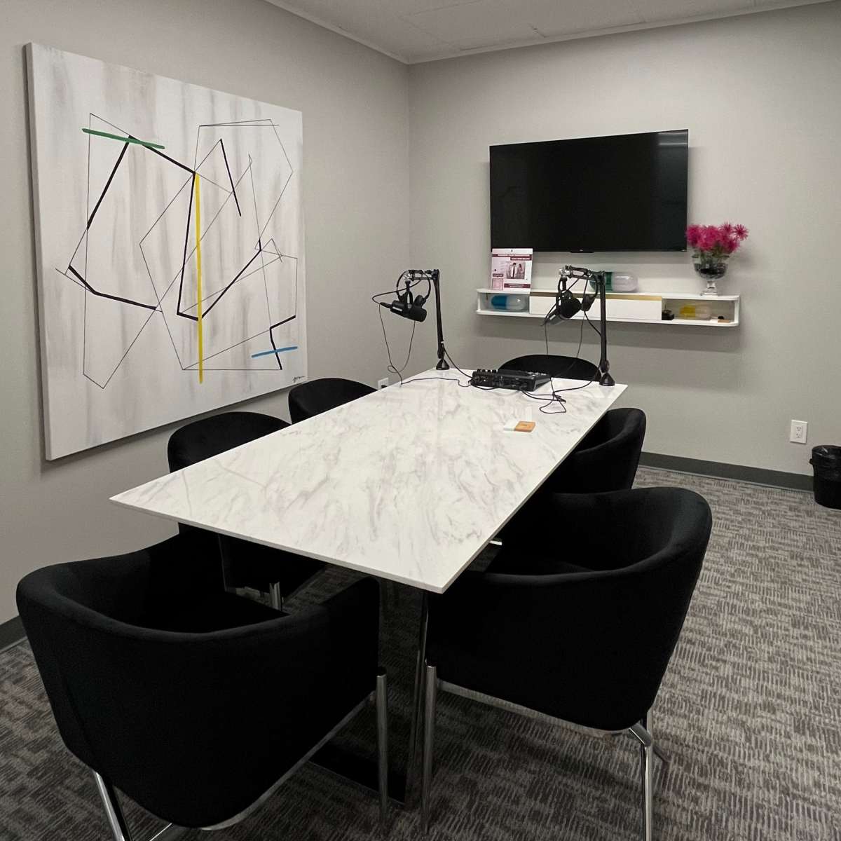 A modern conference room features a marble table surrounded by black chairs, a mounted television, and a large abstract painting on the wall.