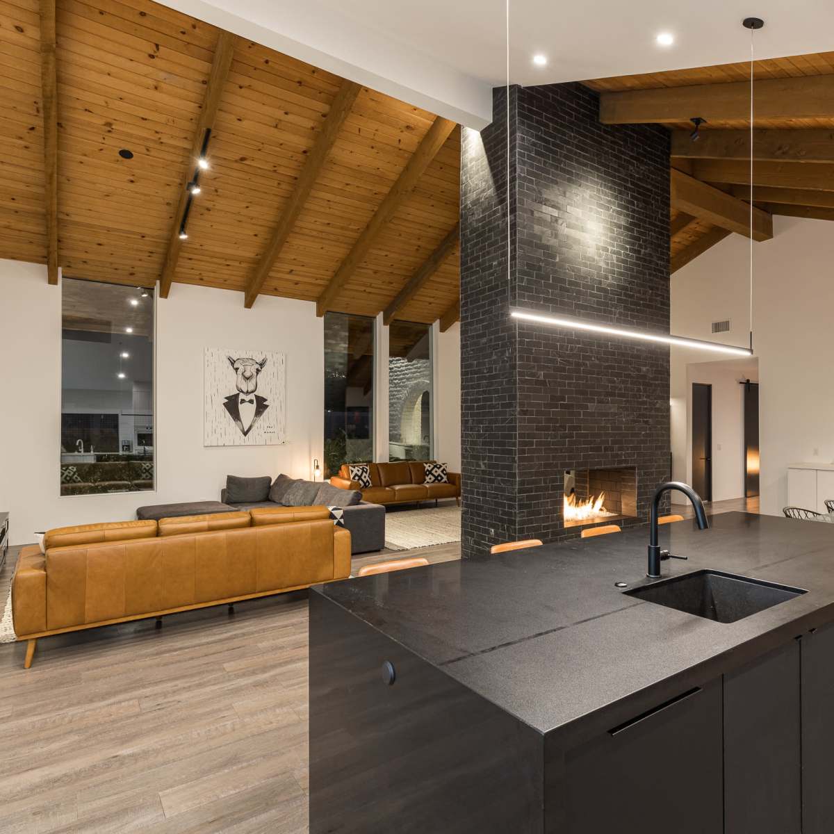 A modern open-concept living space featuring a dark kitchen island, a cozy seating area with a sofa, a large fireplace, and high wooden beam ceilings.