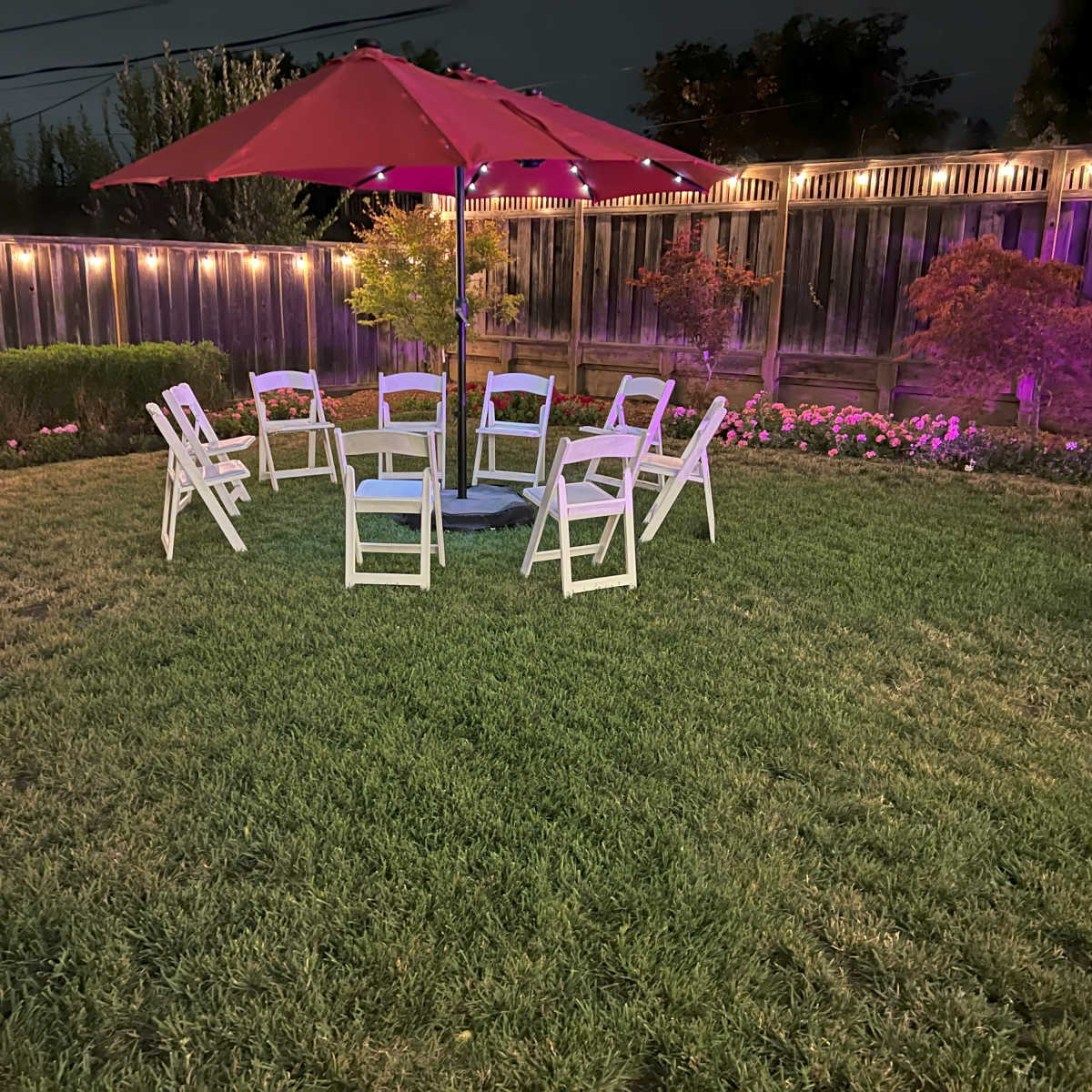 A circular arrangement of white chairs sits on a grassy area beneath a red umbrella, surrounded by blooming flowers and illuminated by string lights in a backyard at dusk.