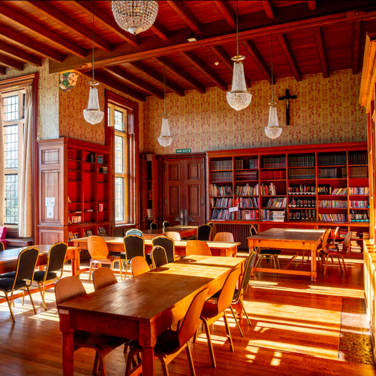 A bright, spacious library with wooden tables and chairs arranged around a room filled with bookshelves and large windows allowing natural light to enter.