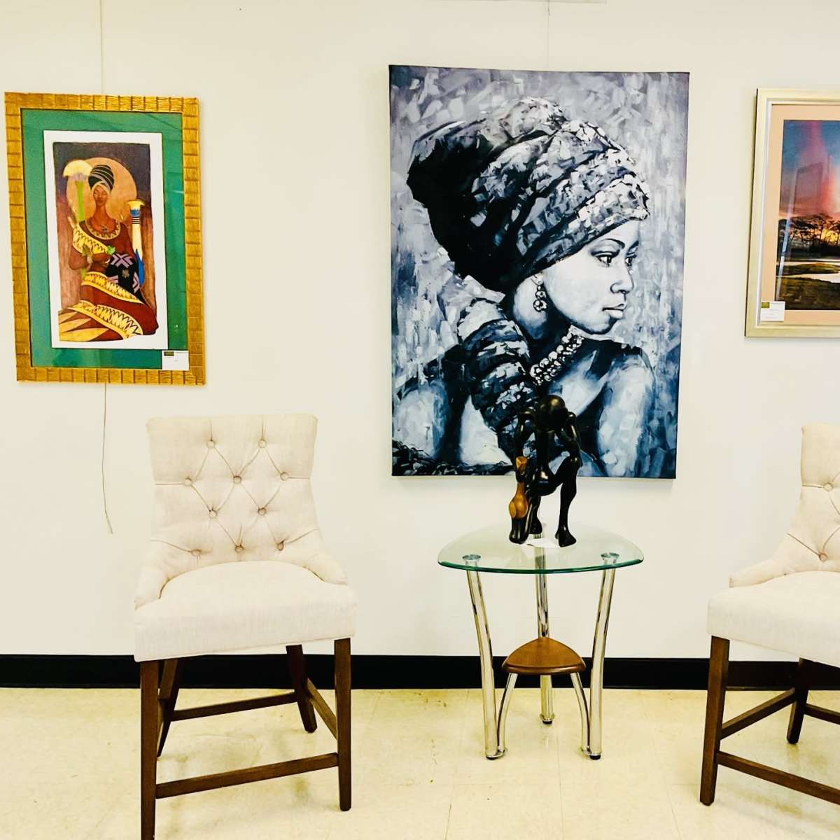 A small seating area with two upholstered chairs, a glass side table featuring a statue, and a diverse collection of framed artwork on the wall.