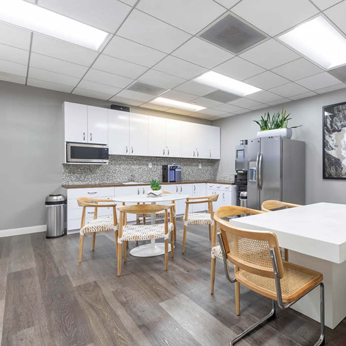 A modern office kitchen with a large white table, wooden chairs, a refrigerator, and a window displaying a coastal view.