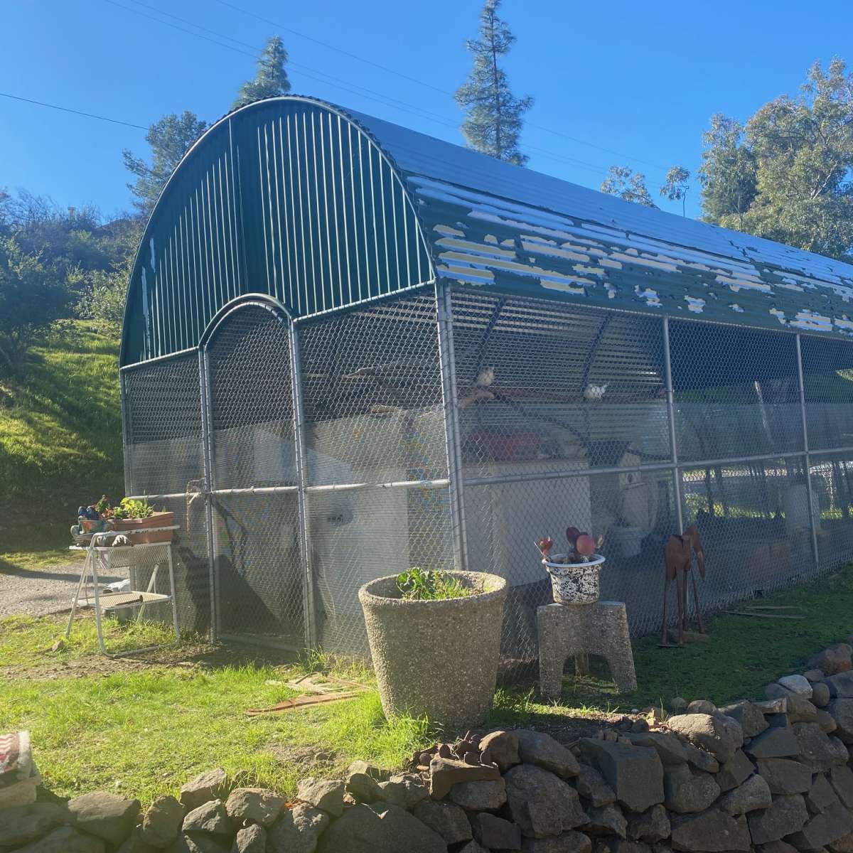 A large metal barn-like structure enclosed by a chain-link fence, situated on a grassy area beside a stone wall.