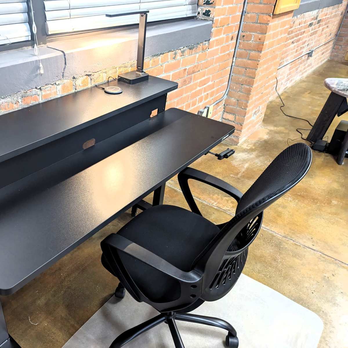 A black desk with a lamp, charging port, and a small potted plant is positioned against a brick wall in a modern workspace.