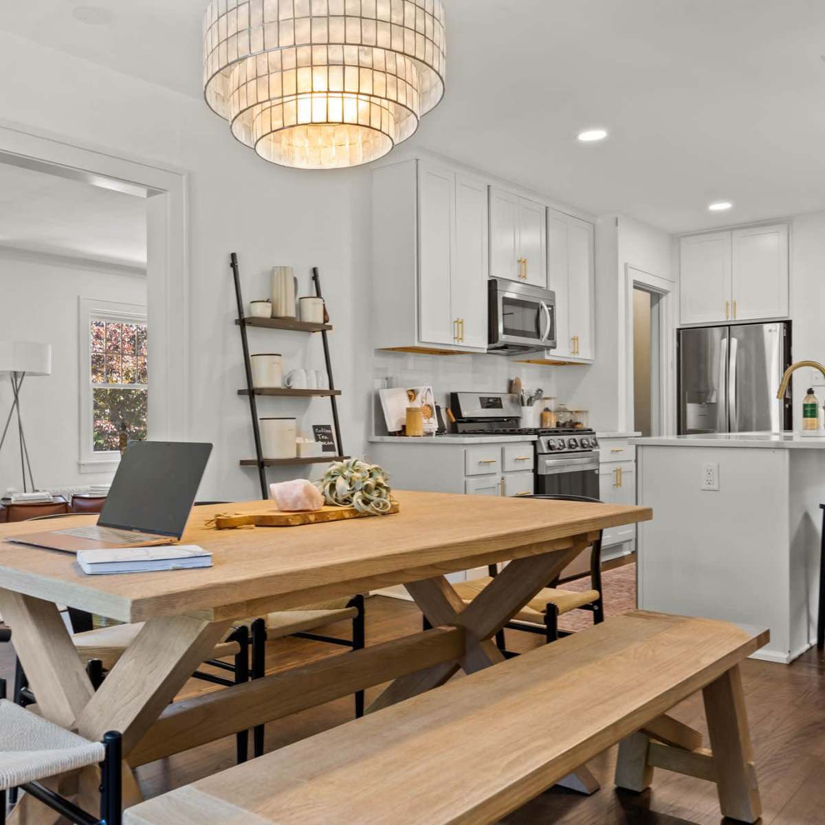 A modern kitchen and dining area featuring a wooden table with benches, a laptop, and a chandelier hanging above.