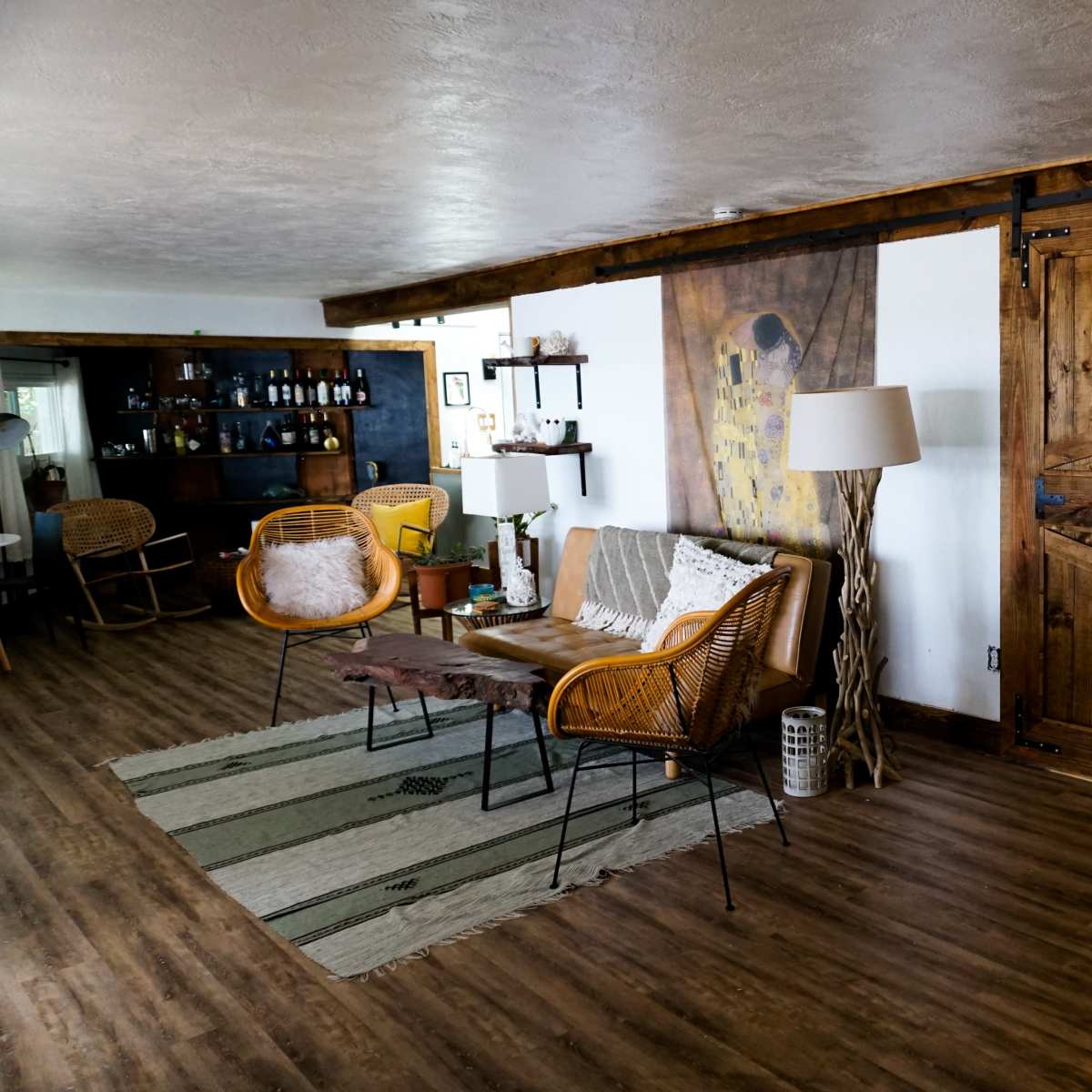 A cozy living room featuring a wooden sliding barn door, a pair of wicker chairs paired with a small table, and a lamp, all set on a wooden floor with a striped rug.