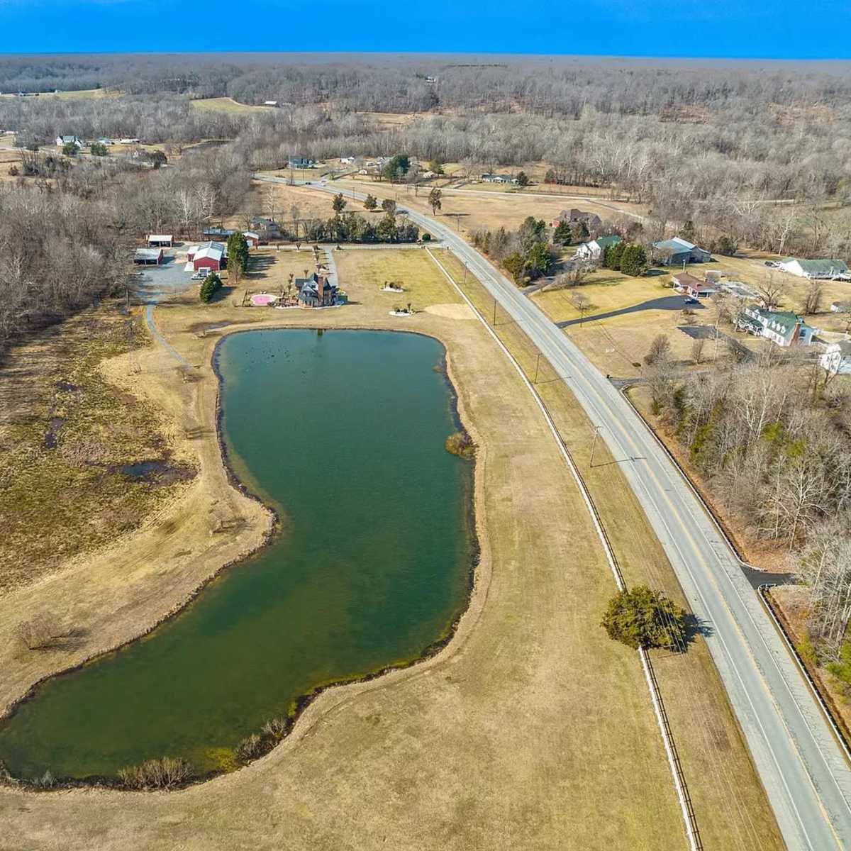 An aerial view of a curved pond next to a road, surrounded by open fields and scattered buildings.