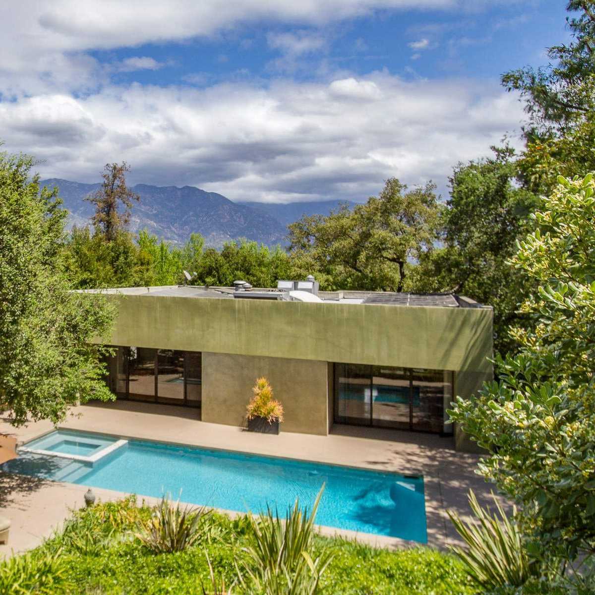 A modern house with large windows beside a swimming pool, surrounded by trees and mountains in the background under a cloudy sky.