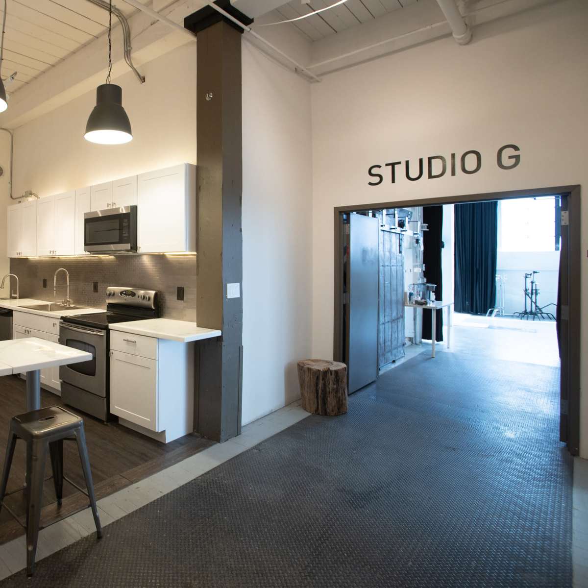 A modern kitchen area adjacent to an entrance labeled "STUDIO G," featuring white cabinetry, stainless steel appliances, and bar seating.