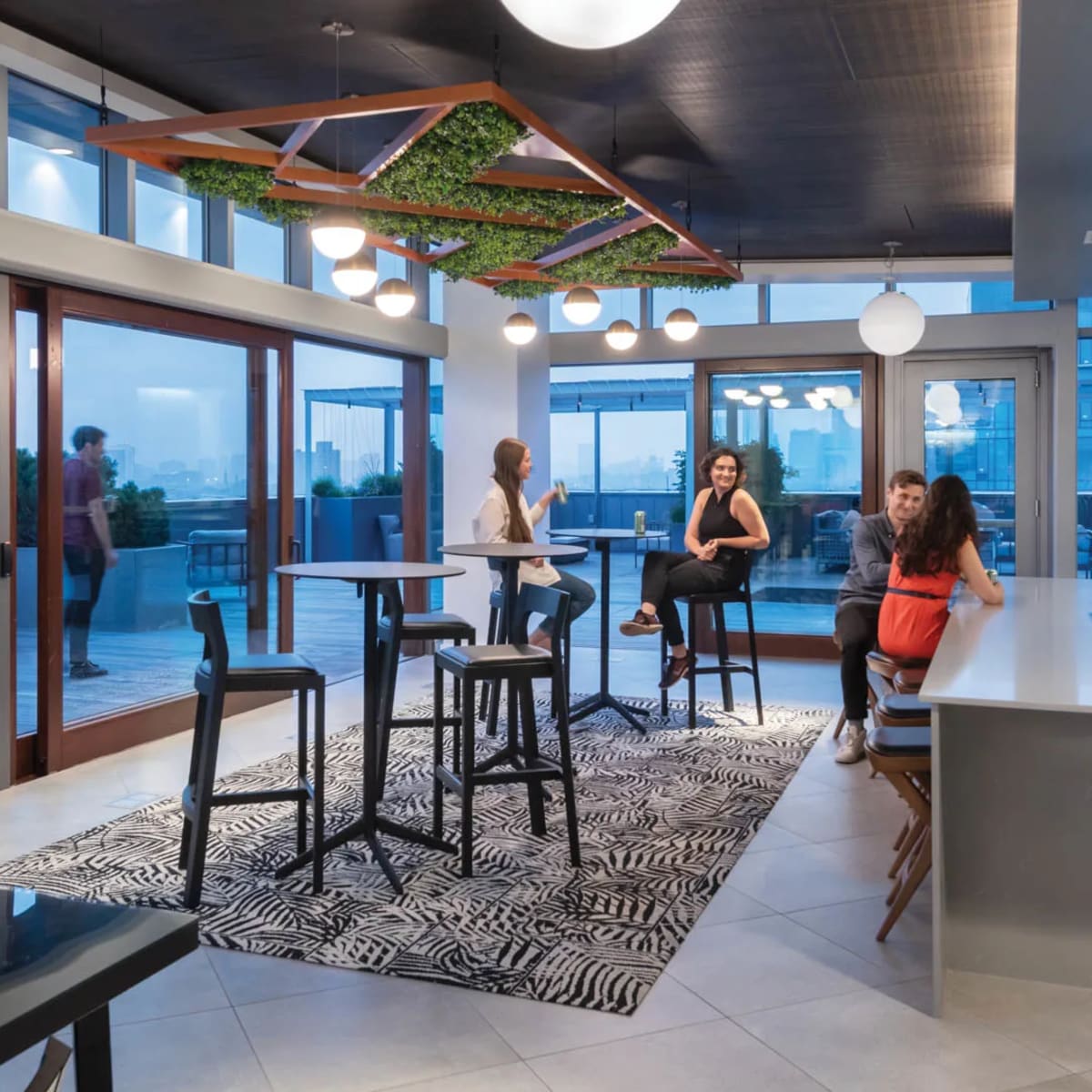A modern lounge area features a kitchen island, bar seating, and tall tables with views of the city through large windows.