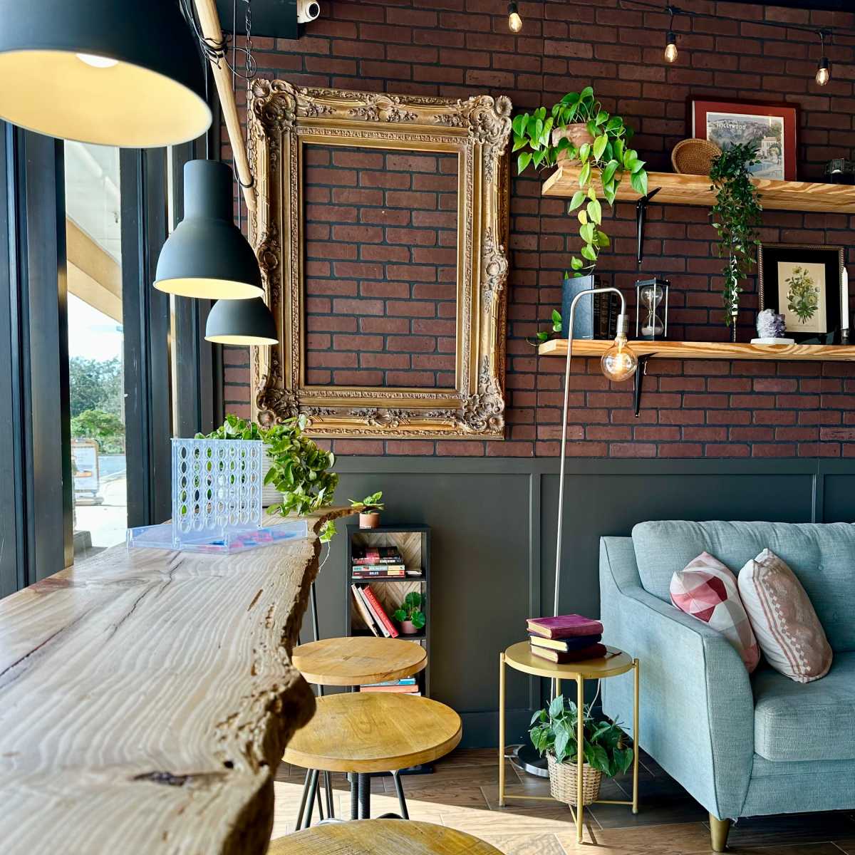 The scene features a cozy café interior with wooden bar stools, a long wooden countertop, a green sofa, and framed artwork along a brick wall adorned with plants and decorative items.