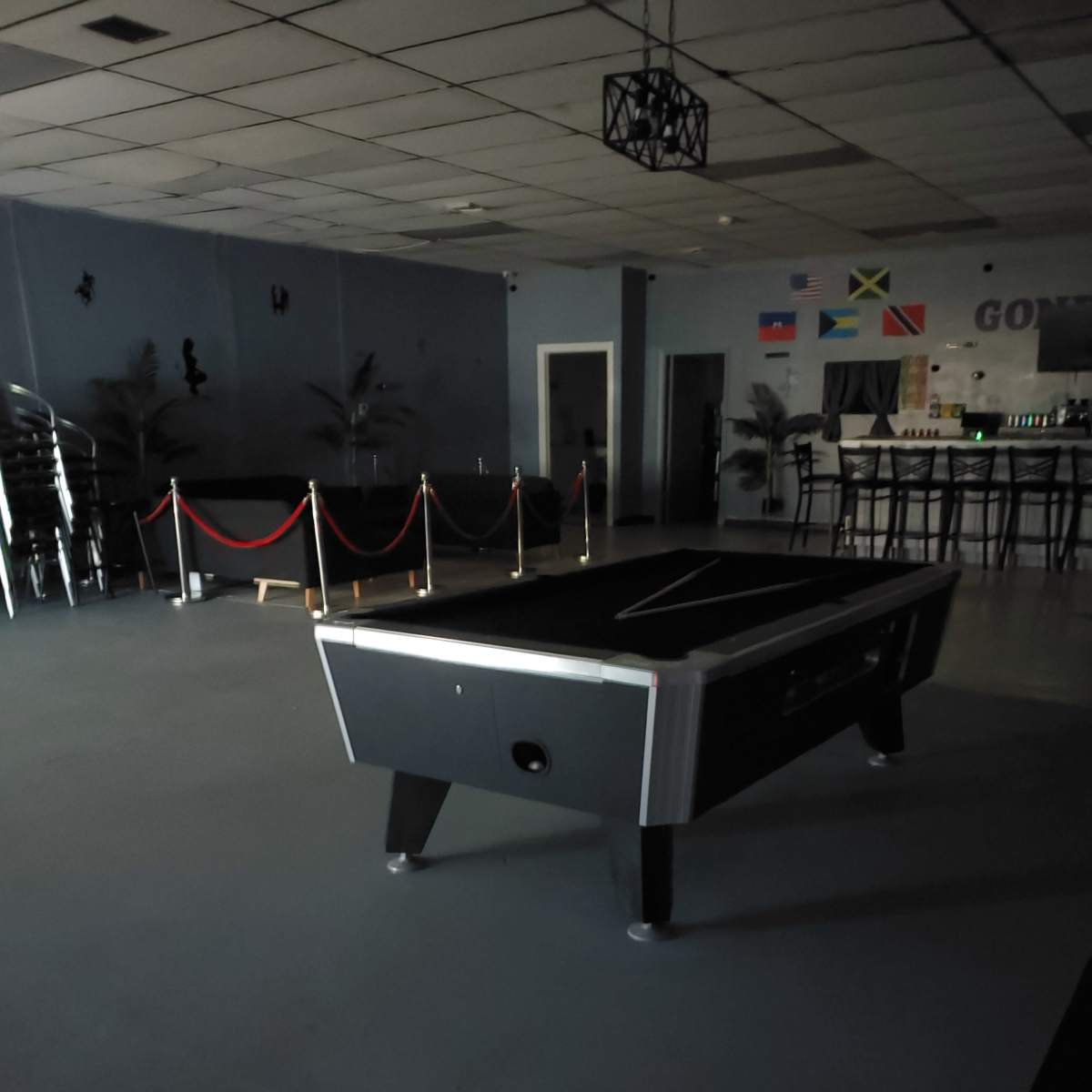 A dimly lit room with a black pool table in the center, surrounded by a red rope barrier and several chairs stacked against the wall.