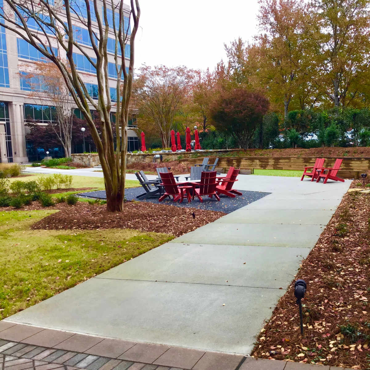 A landscaped outdoor area featuring arranged chairs and tables on a concrete pathway surrounded by grass and trees.