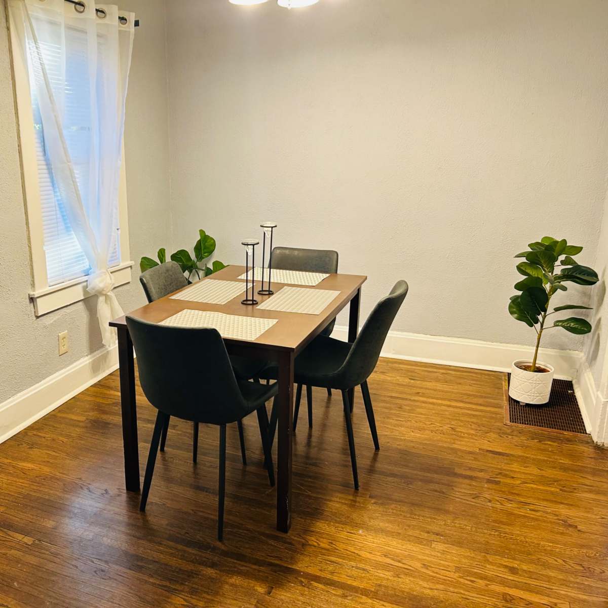 A dining area features a dark wooden table with four chairs, a candle holder on the table, and two potted plants in the corners.