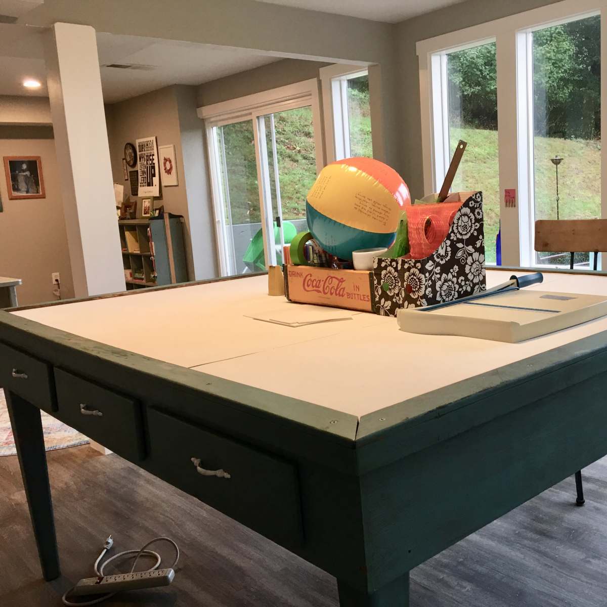 A large, painted teal table with drawers is set in a room filled with various colorful items, including a beach ball and a toolbox, while sunlight streams through the windows.