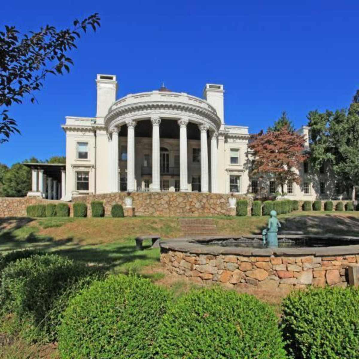 A large mansion with a columned facade, surrounded by neatly trimmed hedges and a circular stone fountain in the foreground.