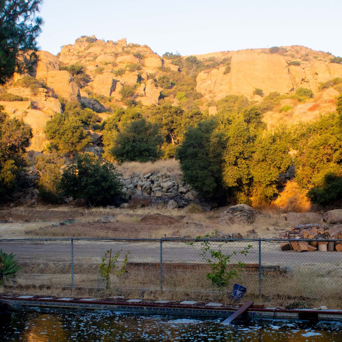 A dry, rocky landscape with a mountainous backdrop and scattered trees, bordered by a fenced area featuring a pool in the foreground.