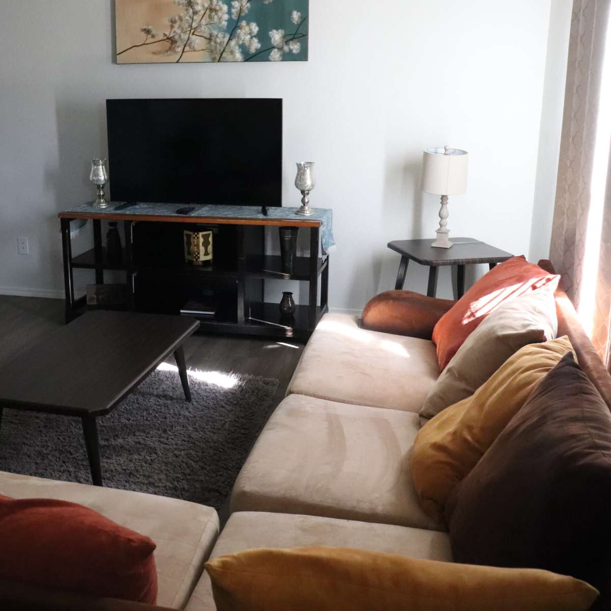 A living room features a sectional sofa, a television on a stand, and a small coffee table, with a doorway leading to another room in the background.