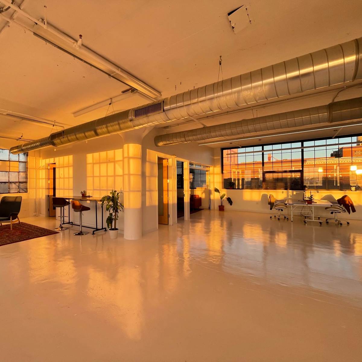 A spacious, well-lit industrial studio features large windows, polished white floors, and minimalist furnishings under soft sunset lighting.