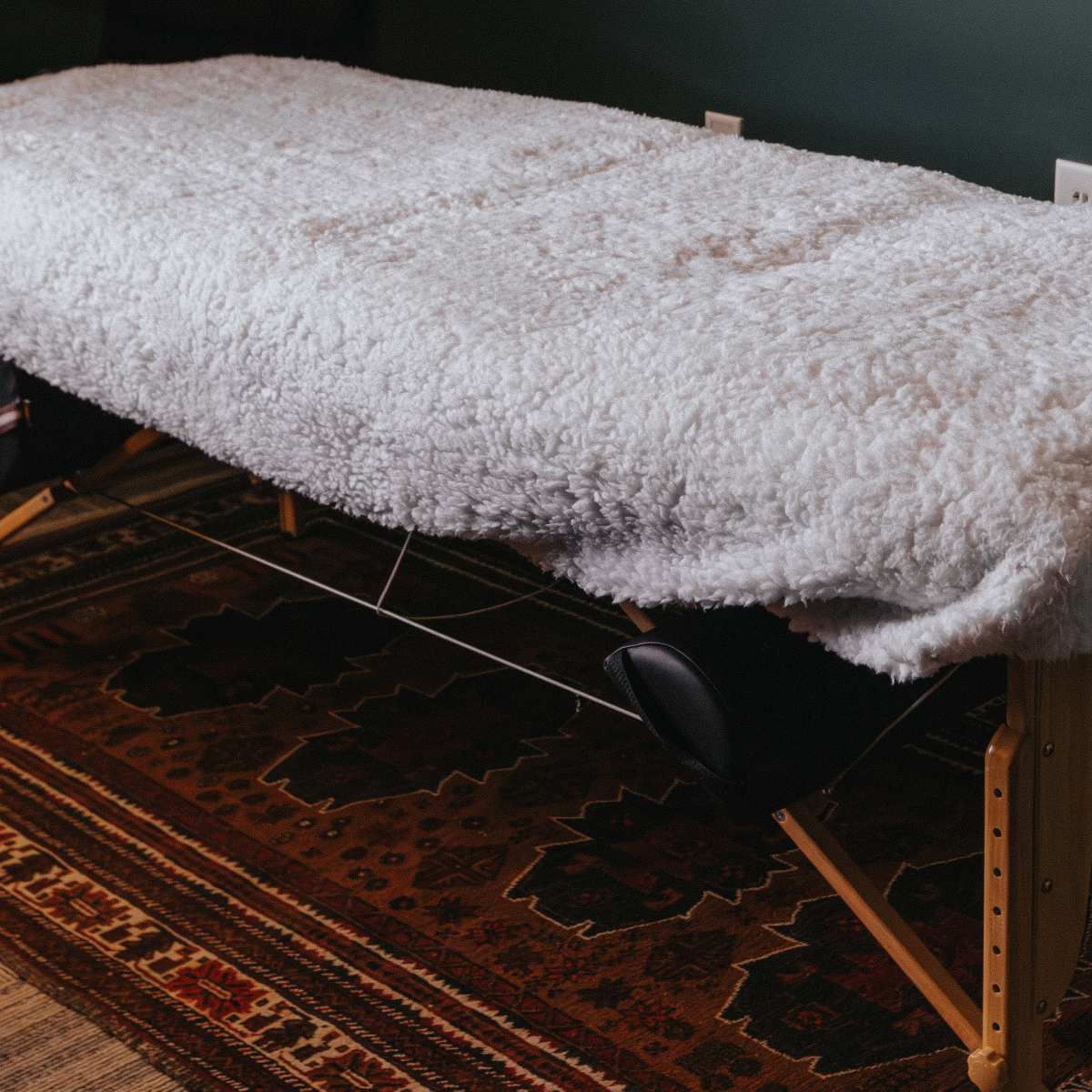 A massage table covered with a white, fluffy blanket sits on a patterned rug in a room with green walls.