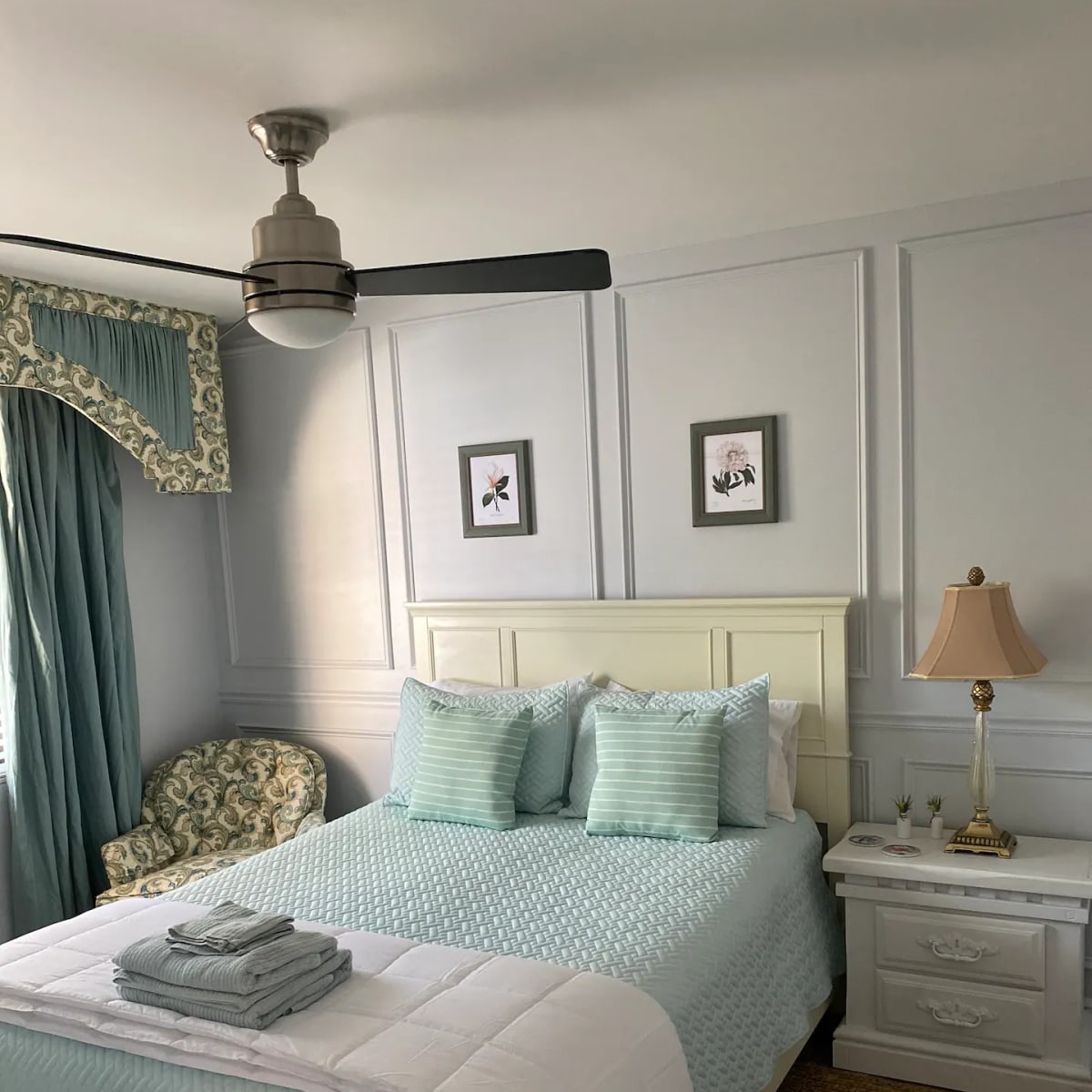 A neatly arranged bedroom featuring a light blue bedspread, decorative pillows, a bedside lamp, and a patterned armchair near a window.