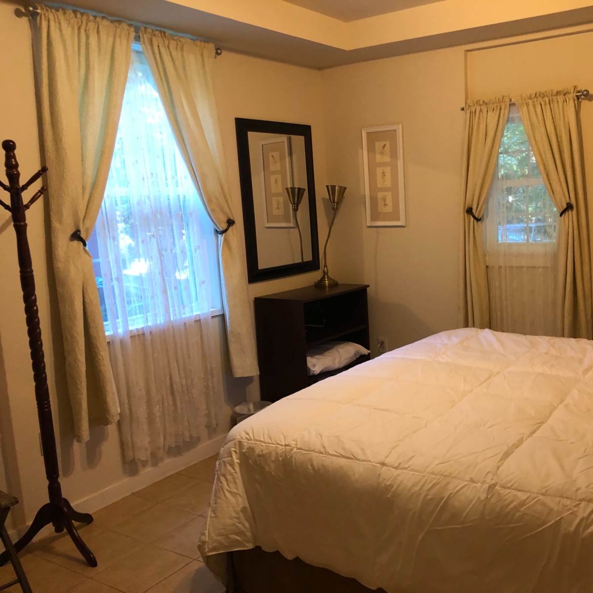 A bedroom with a large bed, light-colored curtains, and a mirror on the wall, beside a window that lets in natural light.