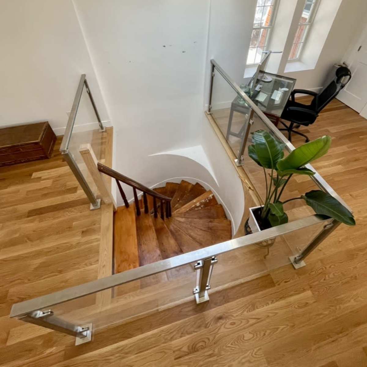 A spiral staircase leading down, surrounded by a glass railing, with a small desk and chair visible in the room at the bottom.