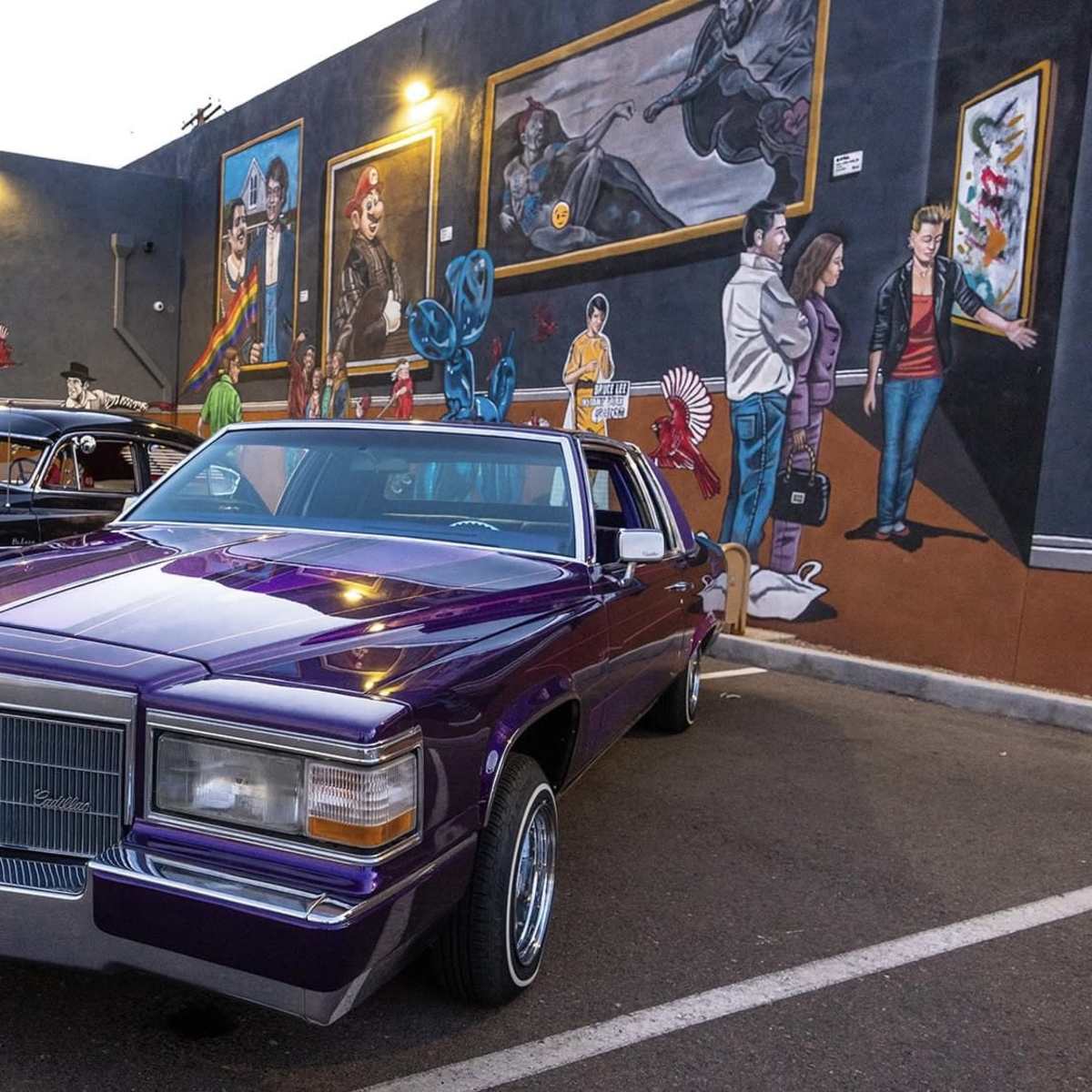 A purple vintage Cadillac is parked in front of a building adorned with colorful murals, alongside two other classic cars.