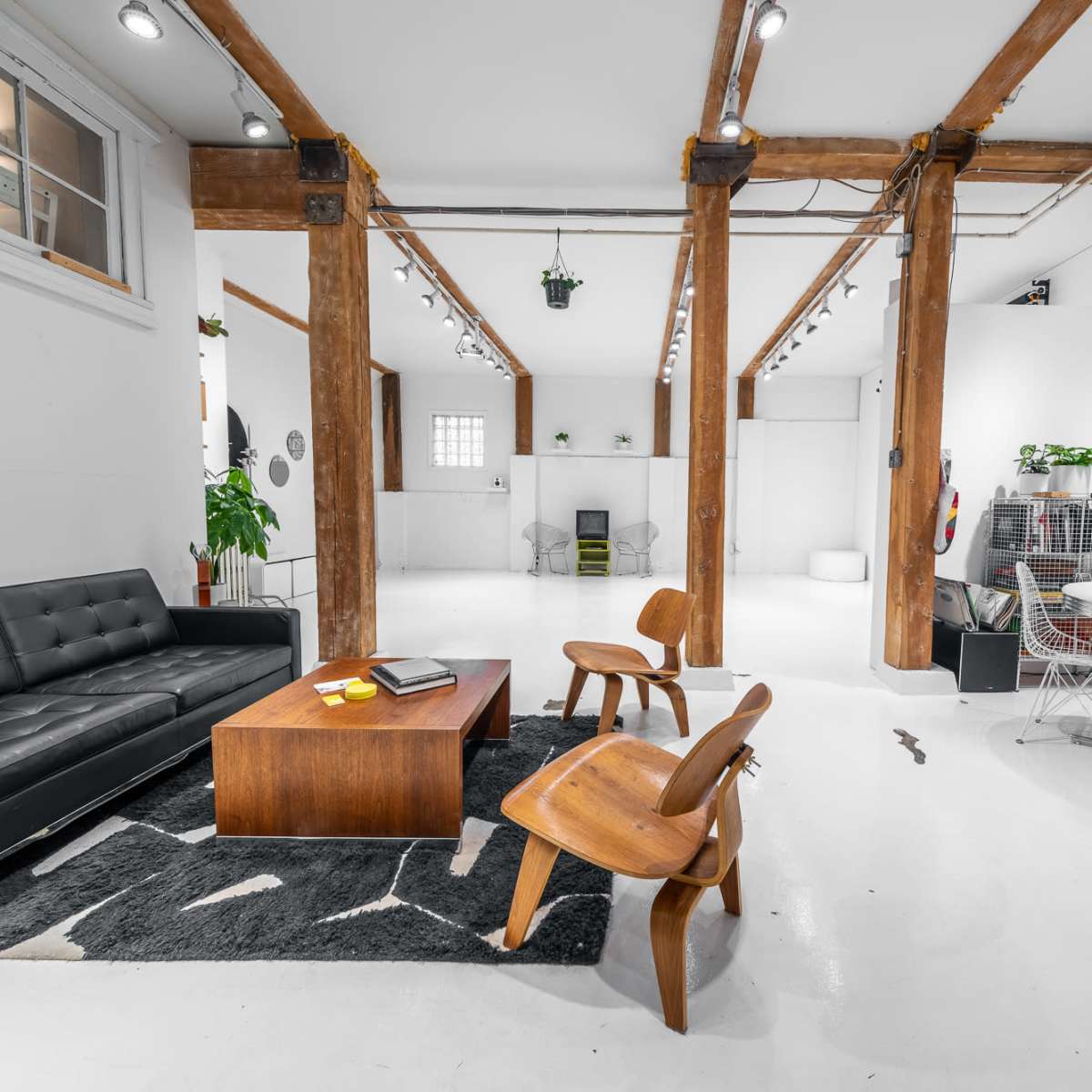 A spacious, industrial-style room featuring a black leather couch, wooden chairs, a coffee table, and various plants.