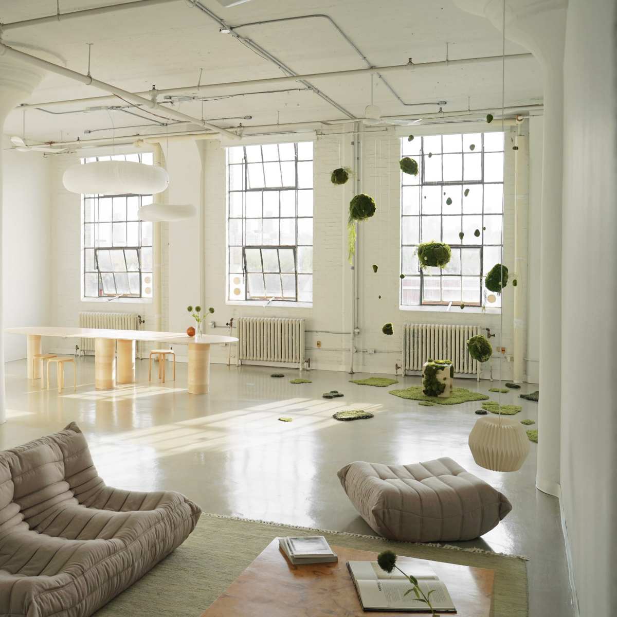 A minimalist interior space with large windows, featuring light-colored walls, various seating arrangements, and hanging green decor.