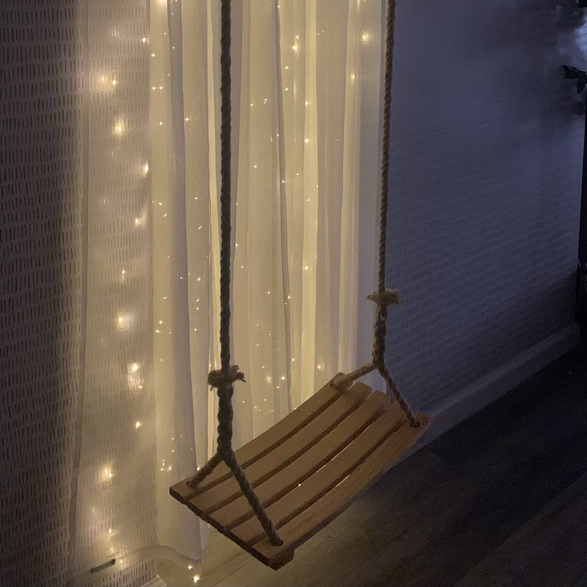 A wooden swing hangs from the ceiling in front of a wall adorned with string lights.