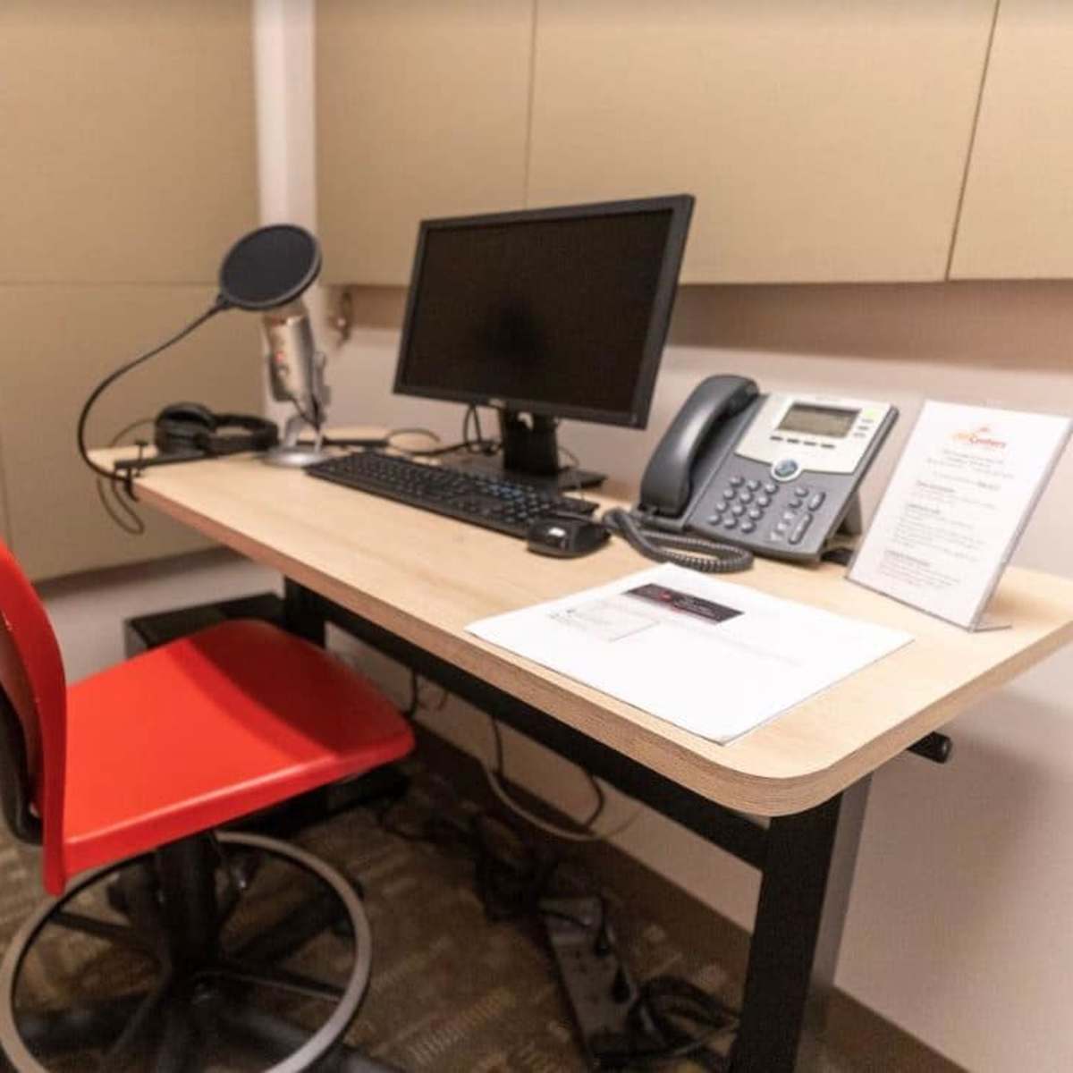 A small office space with a black desk, a computer monitor, a microphone, a phone, and a red chair.
