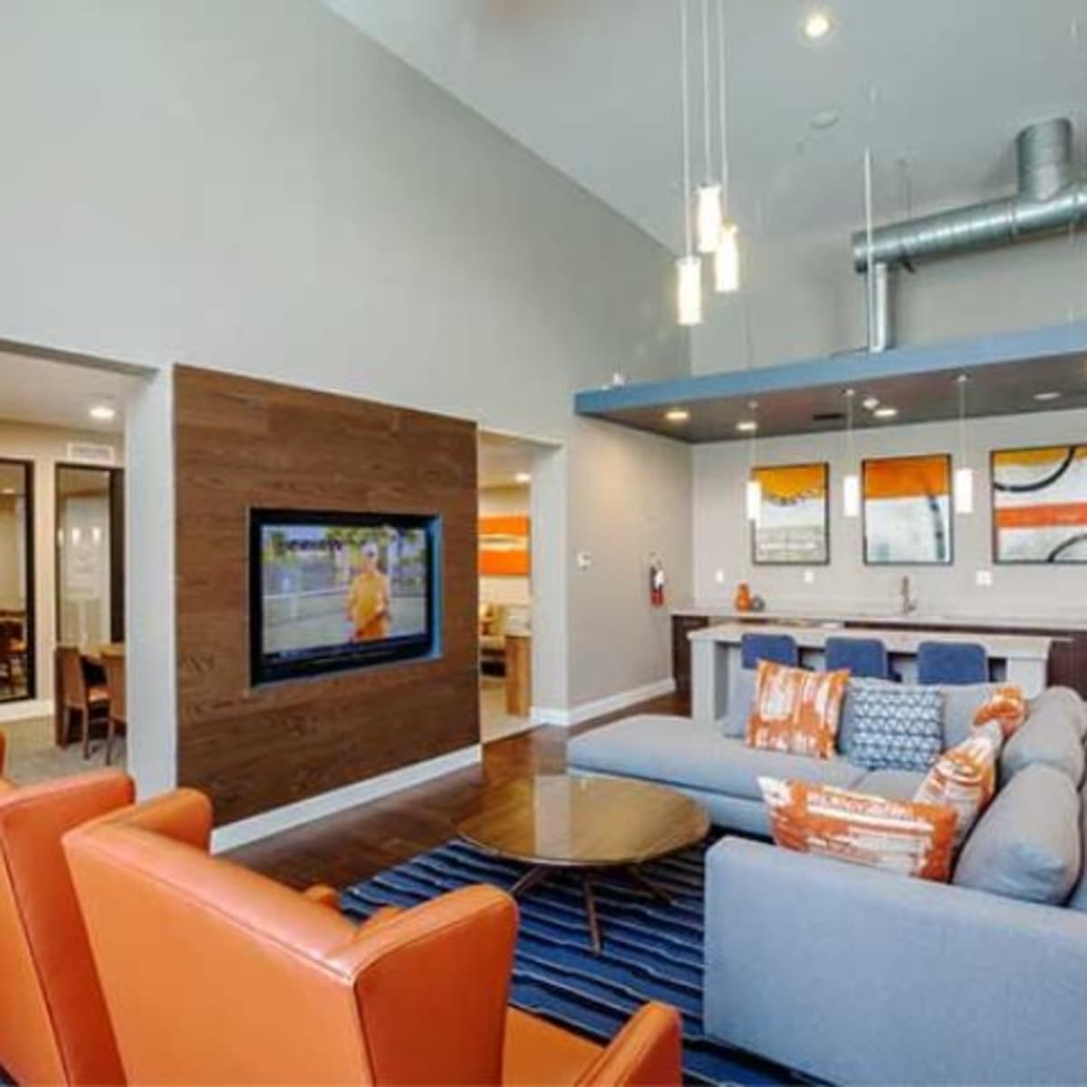 A modern common area with a living room featuring orange and blue furnishings, a TV on the wall, and a dining area in the background.
