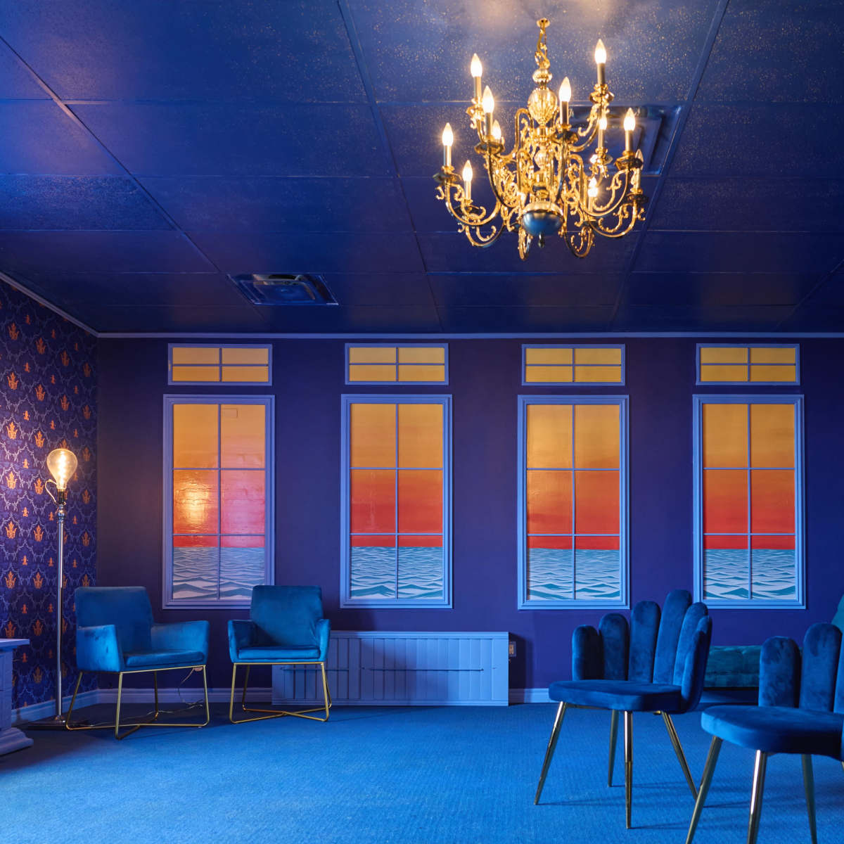 A room with blue walls adorned with patterned wallpaper, featuring a chandelier and several blue chairs arranged around a carpeted floor.