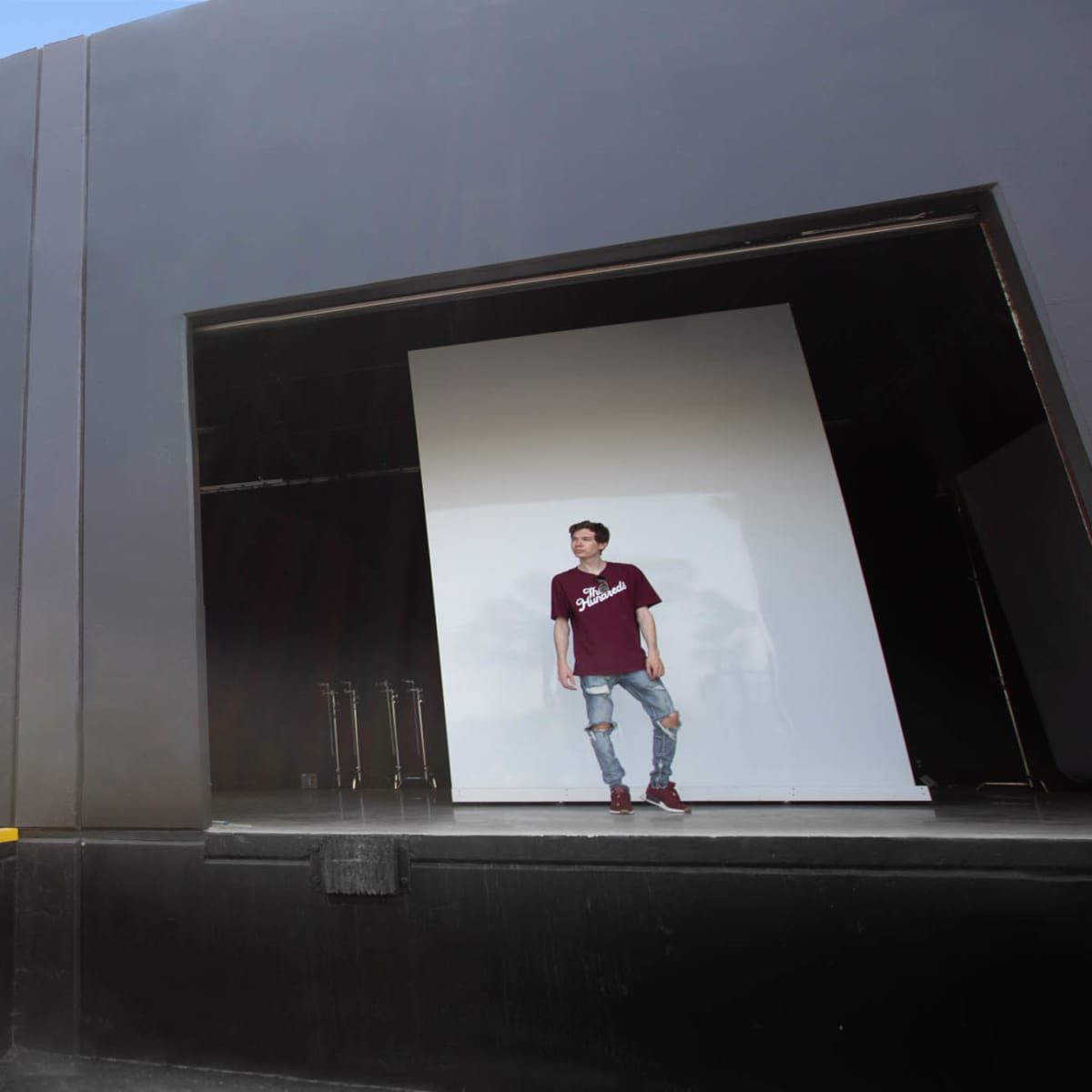 A person stands outside a building with two large openings, one filled with a blank wall and the other revealing an interior space.