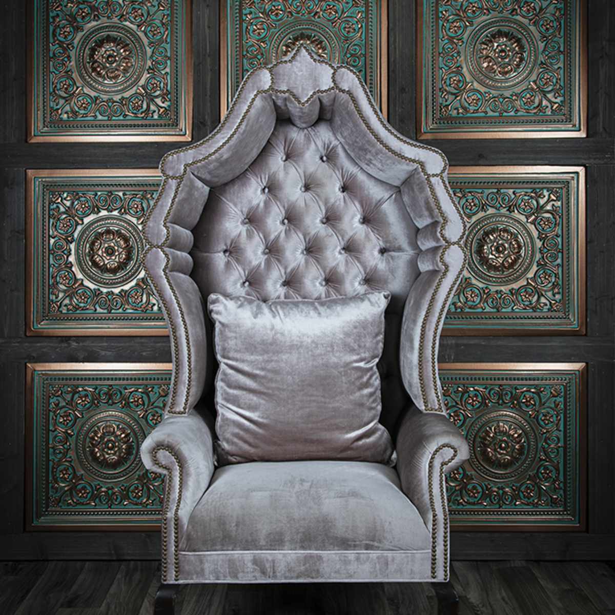 An ornate, gray velvet tufted armchair against a dark wood-paneled wall with intricate teal and gold detailing.