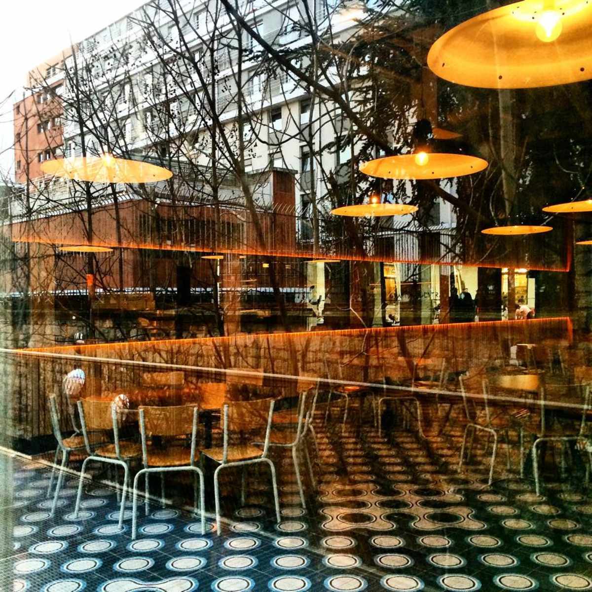A café interior featuring empty chairs and tables, with reflections of trees and buildings visible through the large window.