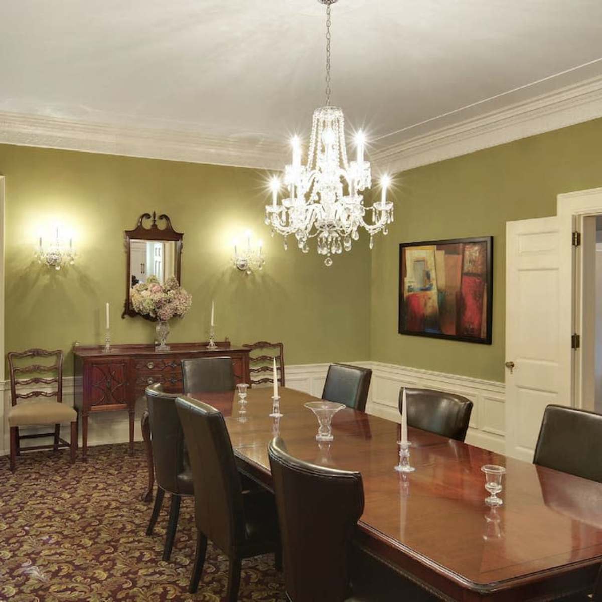 A well-furnished dining room featuring a long wooden table, chairs, a chandelier, and a sideboard against a green wall.