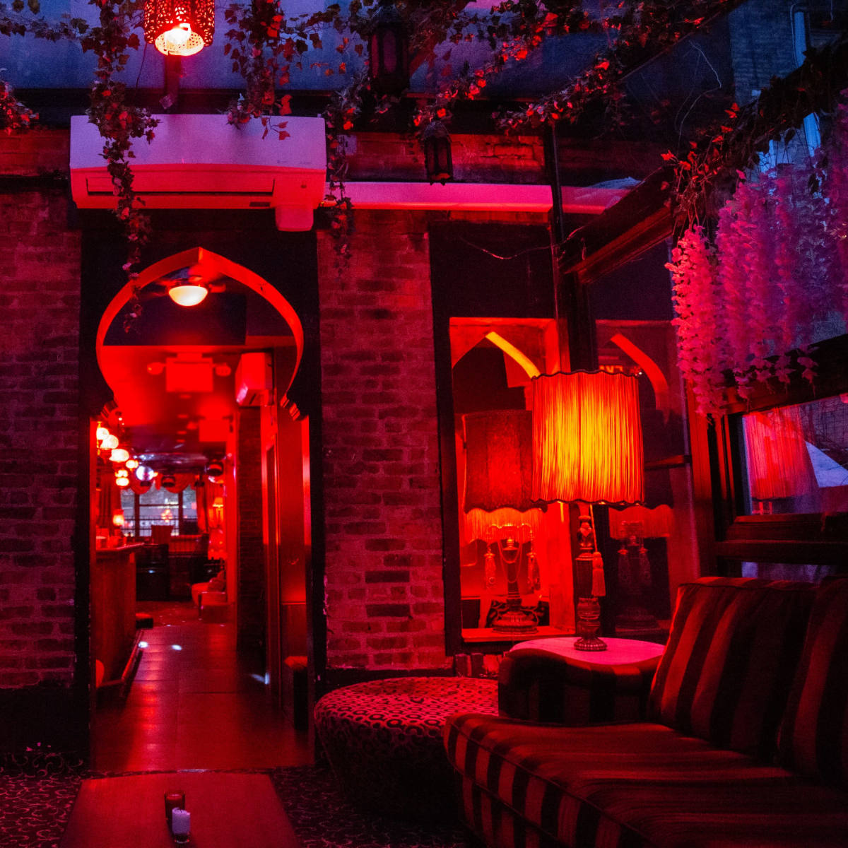 A dimly lit lounge with red lighting, brick walls, and patterned furnishings, featuring lamps and decorative plants.