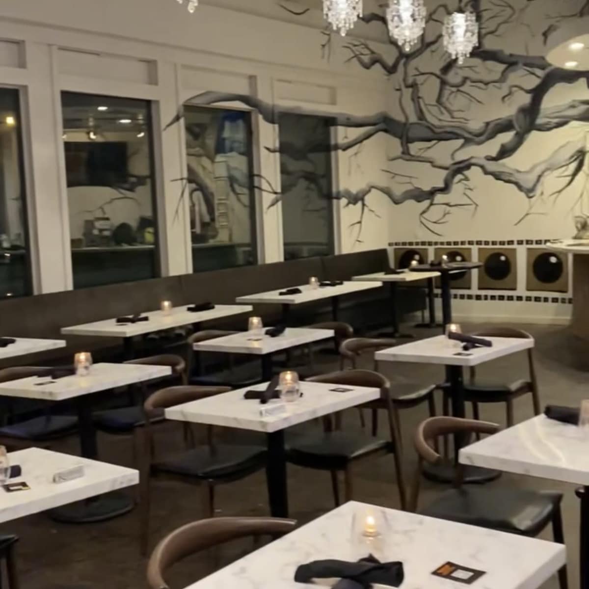 A modern restaurant interior with marble-topped tables, dark chairs, and a wall adorned with an artistic tree mural, illuminated by hanging chandeliers.