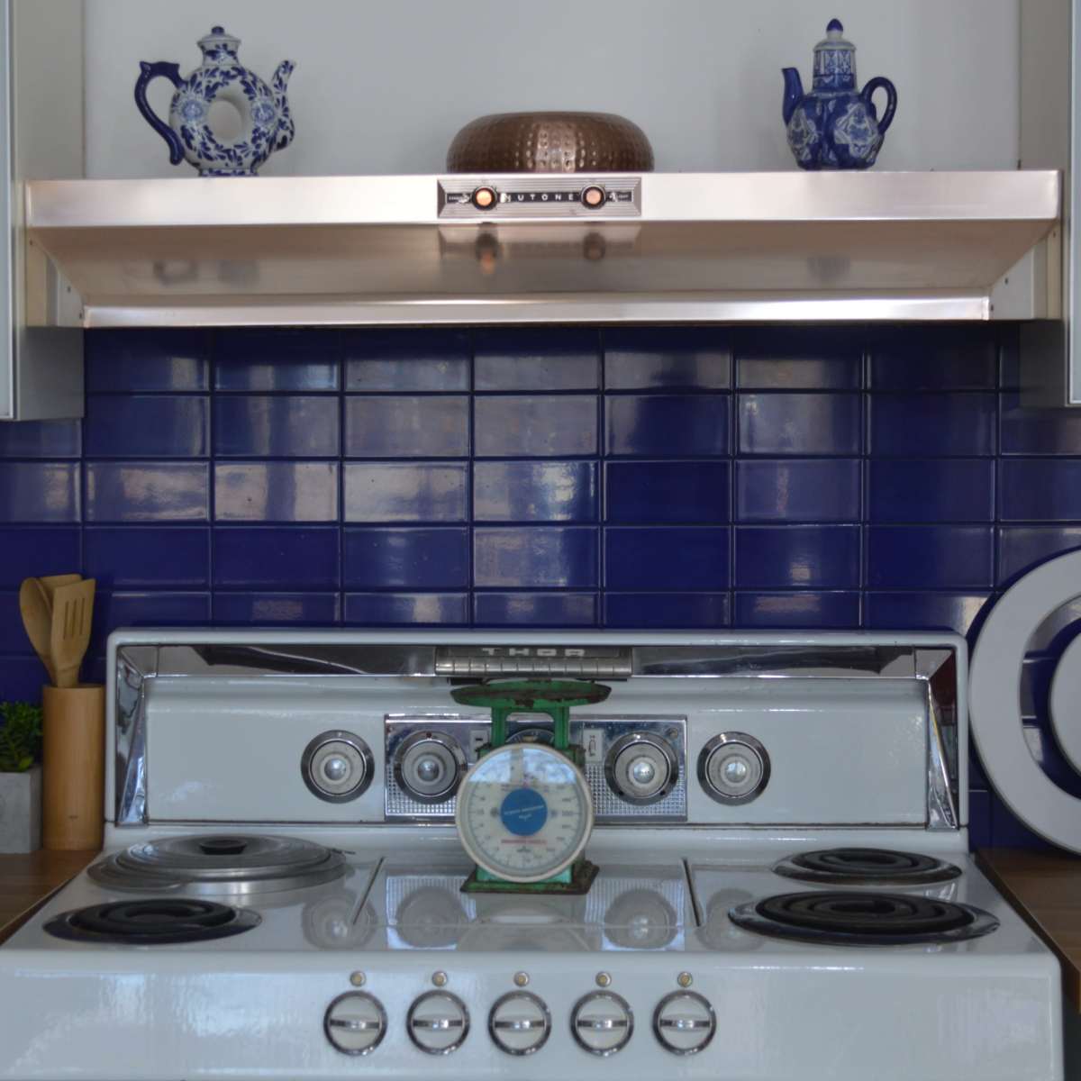A kitchen stove with a green scale on top and blue tiled walls, adorned with decorative items on the shelf above.