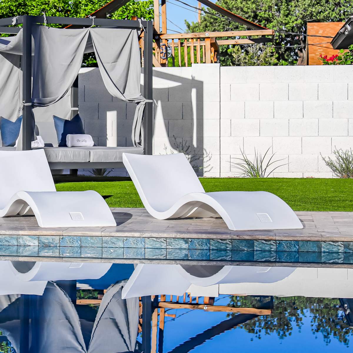 Two white lounge chairs beside a swimming pool, with a canopy-covered seating area in the background and green grass surrounding the setting.
