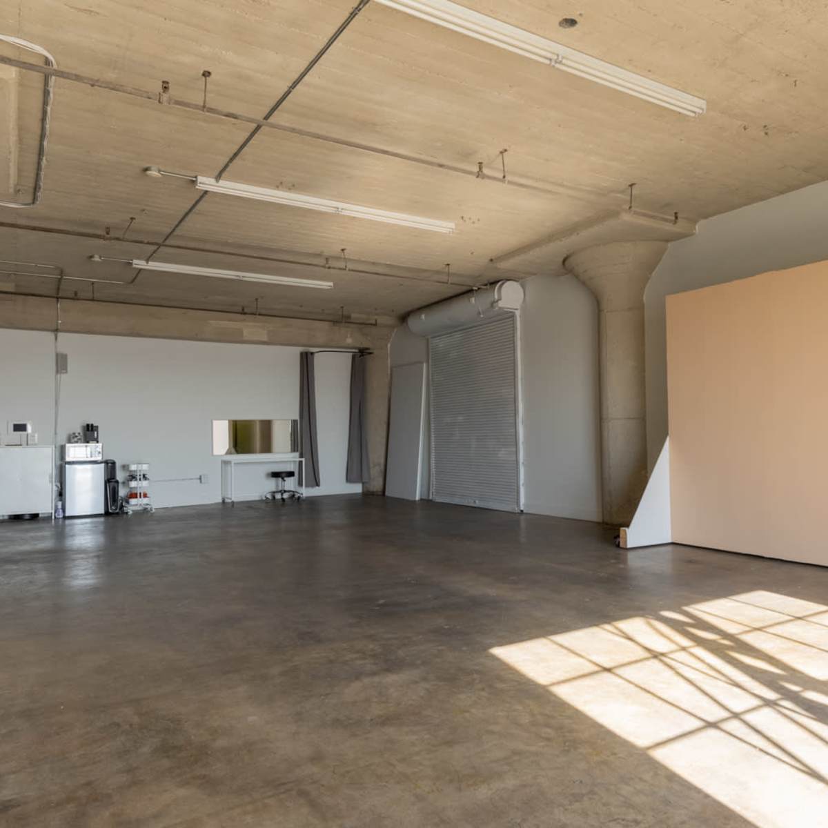 A spacious, empty studio with concrete floors, a large blank backdrop, and a kitchenette area in the background.
