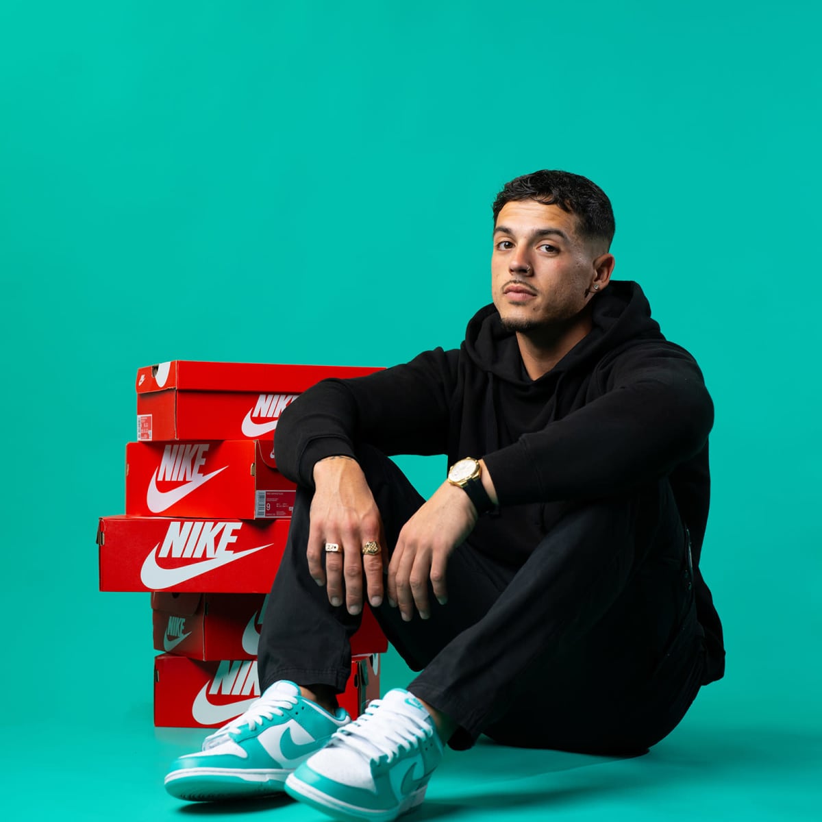 A man sits in front of a stack of red Nike shoeboxes, wearing a black hoodie and light blue sneakers, against a vibrant teal background.