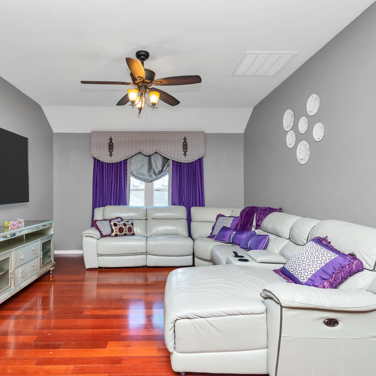 A modern living room features a large flat-screen TV, a light-colored leather sectional sofa, and purple accents on the curtains and throw pillows against gray walls.