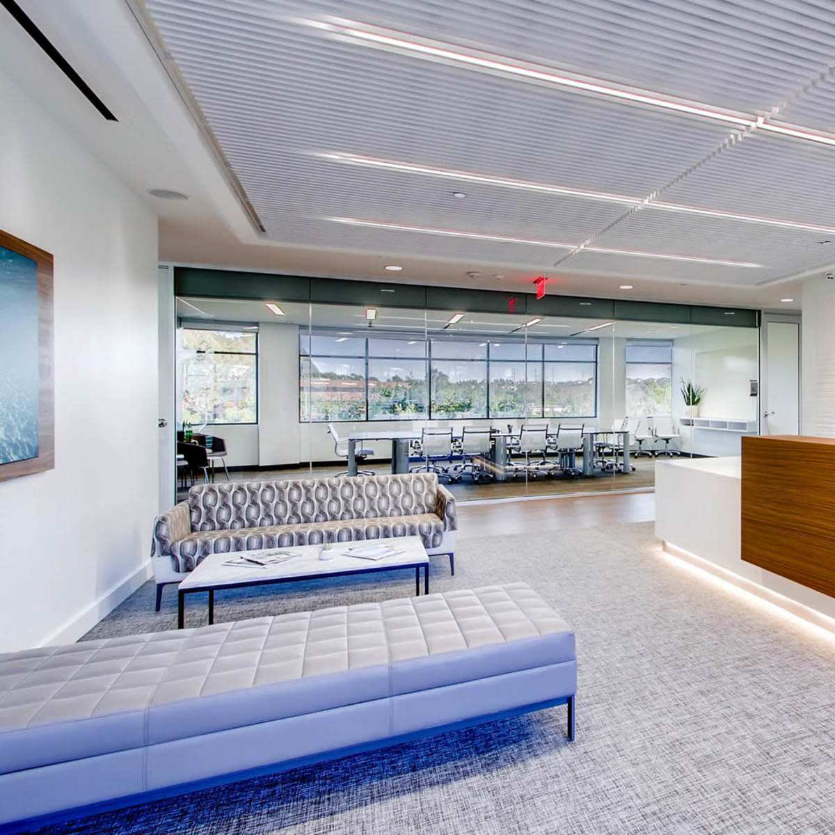 A modern office reception area with a wooden front desk, seating arrangements, and a conference room visible through large windows.