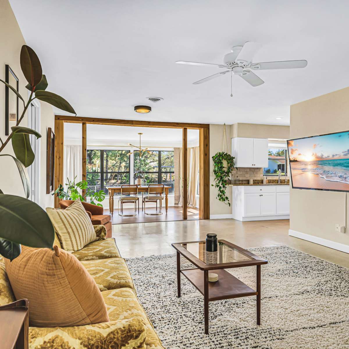 A living room features a sofa, a coffee table, and a mounted television, leading to a dining area with large windows that open to a view of greenery.