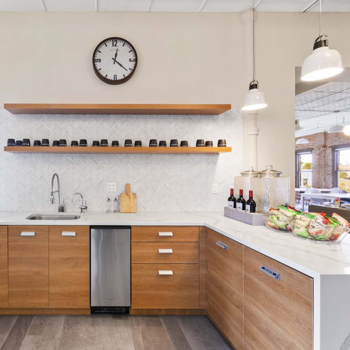 A modern kitchen featuring wooden cabinetry, a sink, and a clock mounted on the wall, with wine bottles and snacks displayed on a shelf.
