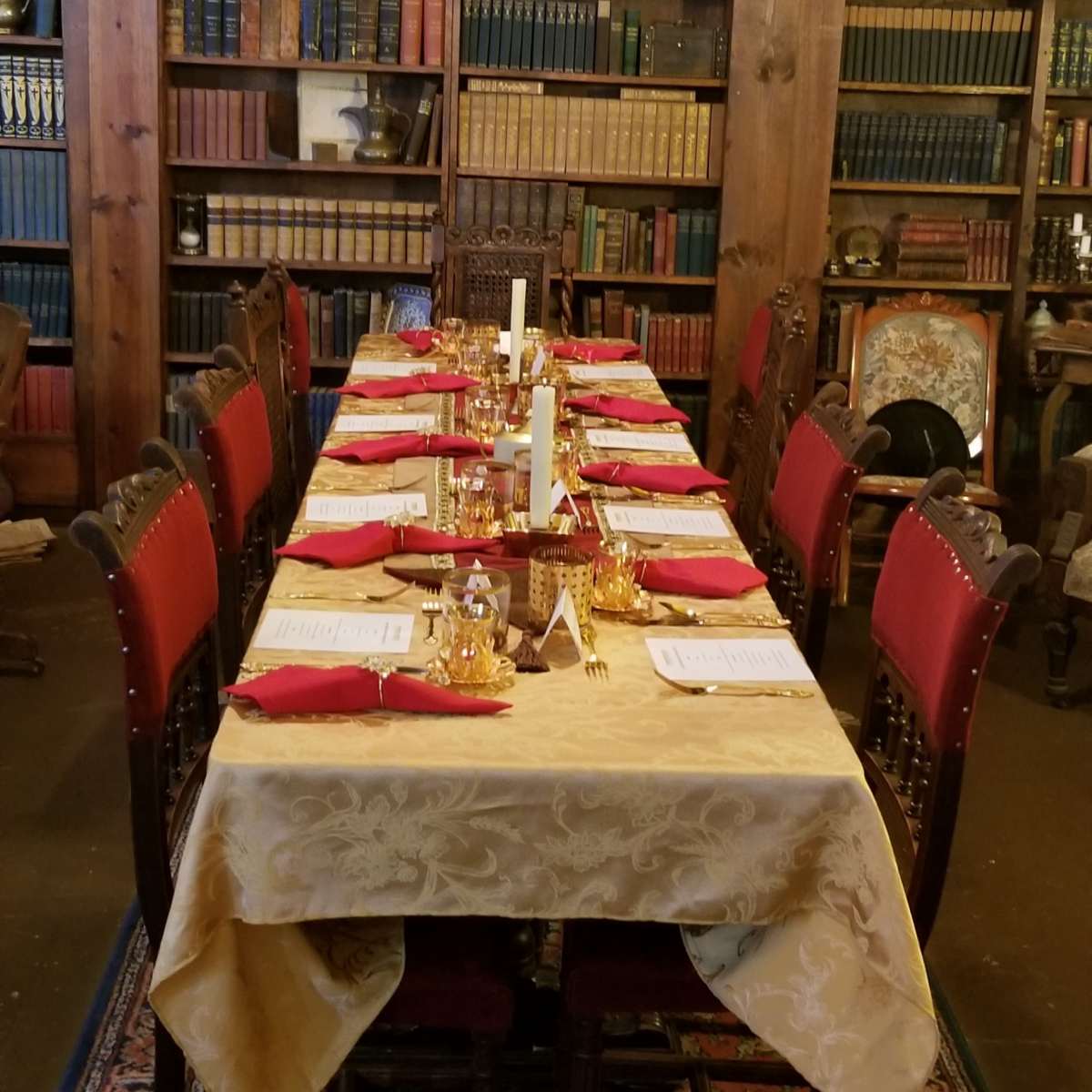 A long dining table is set with red napkins and gold accents in front of wooden bookshelves filled with books.